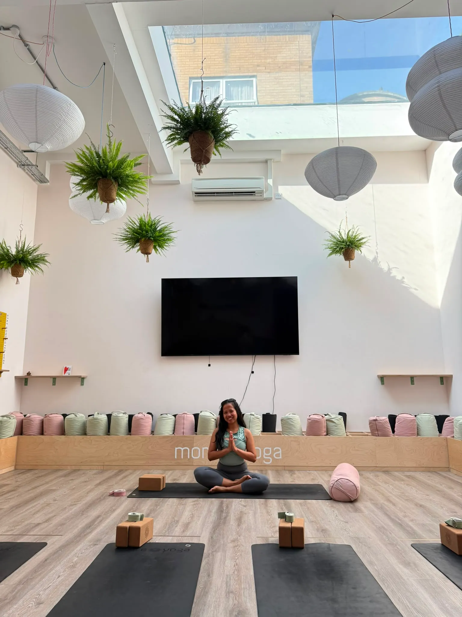 Ardita sitting cross-legged on a yoga mat, hands at heart centre, smiling in a bright, plant-filled studio