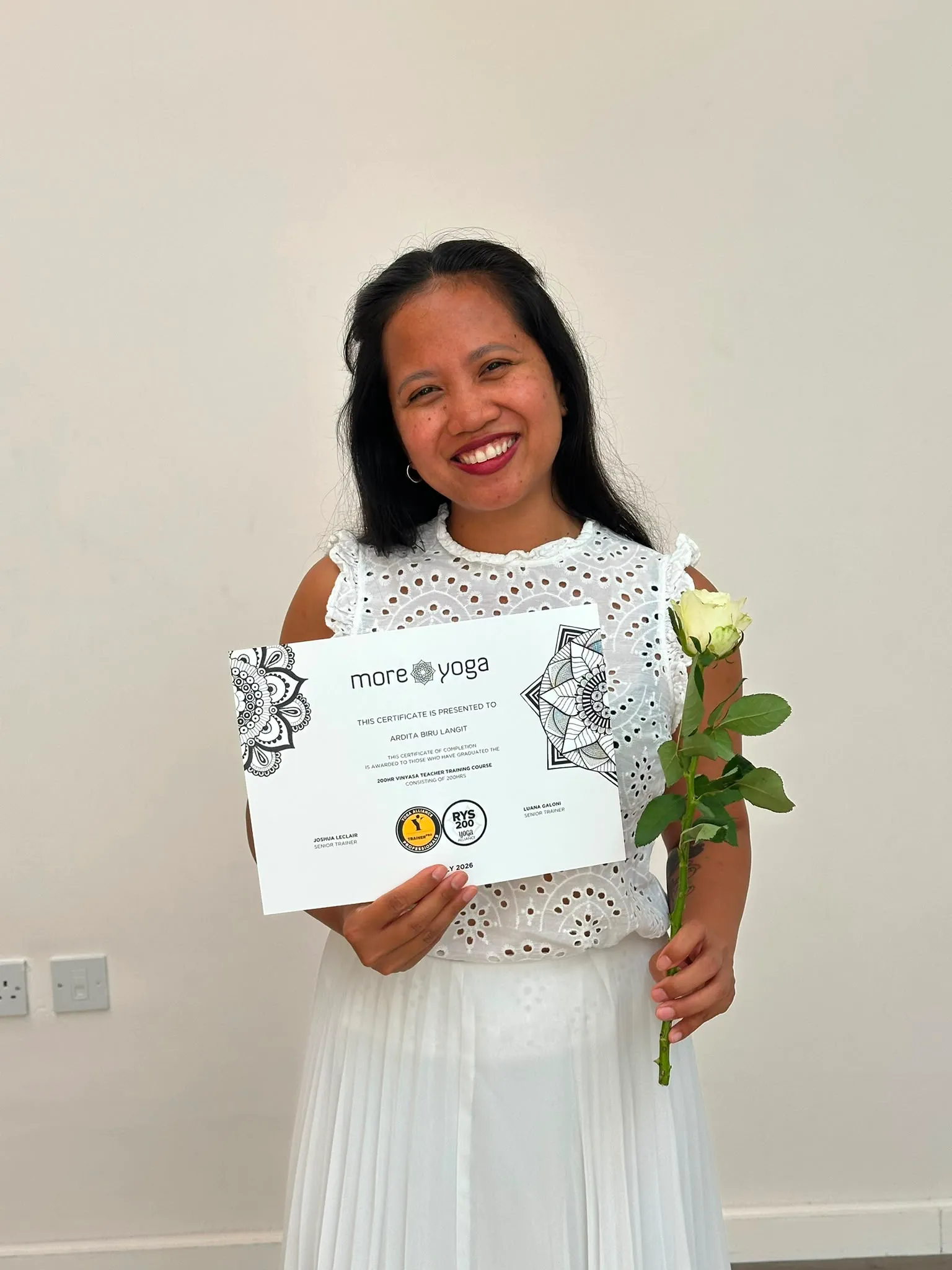 Ardita smiling in white, holding her 200hr vinyasa teacher-training certificate and a white rose